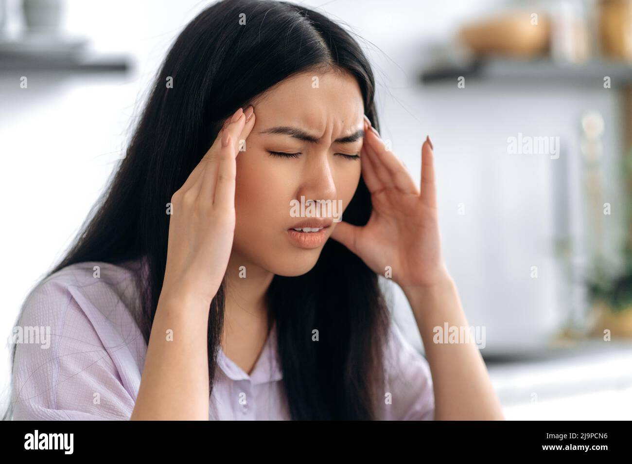Overworked sad chinese brunette girl, tired of working at a laptop, or experiencing a cold, sitting in the kitchen, suffering of headache, massaging her temples, closing eyes, needs rest and treatment Stock Photo