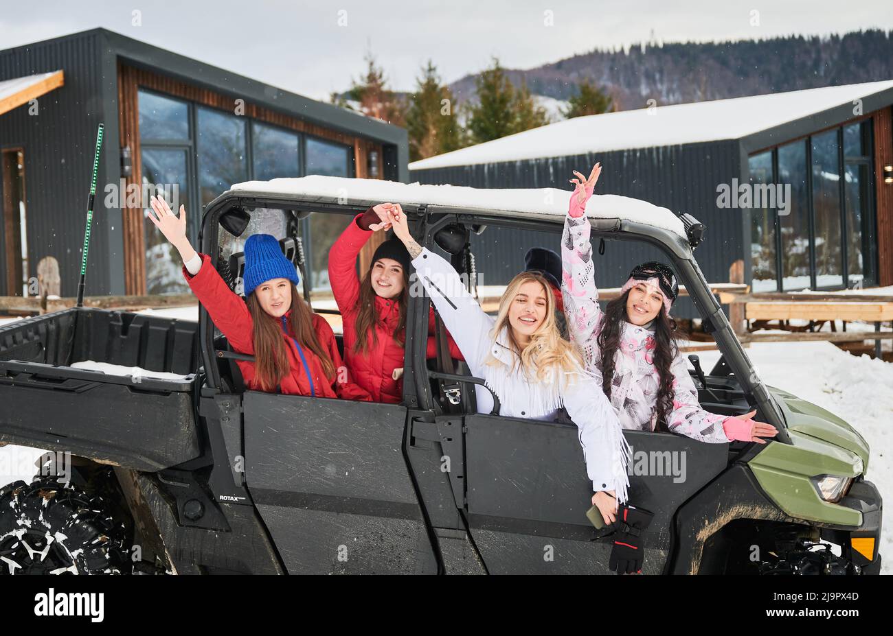 Happy women riding on off-road buggy car. Contemporary barnhouse on background. Concept of active leisure and winter activities. Stock Photo