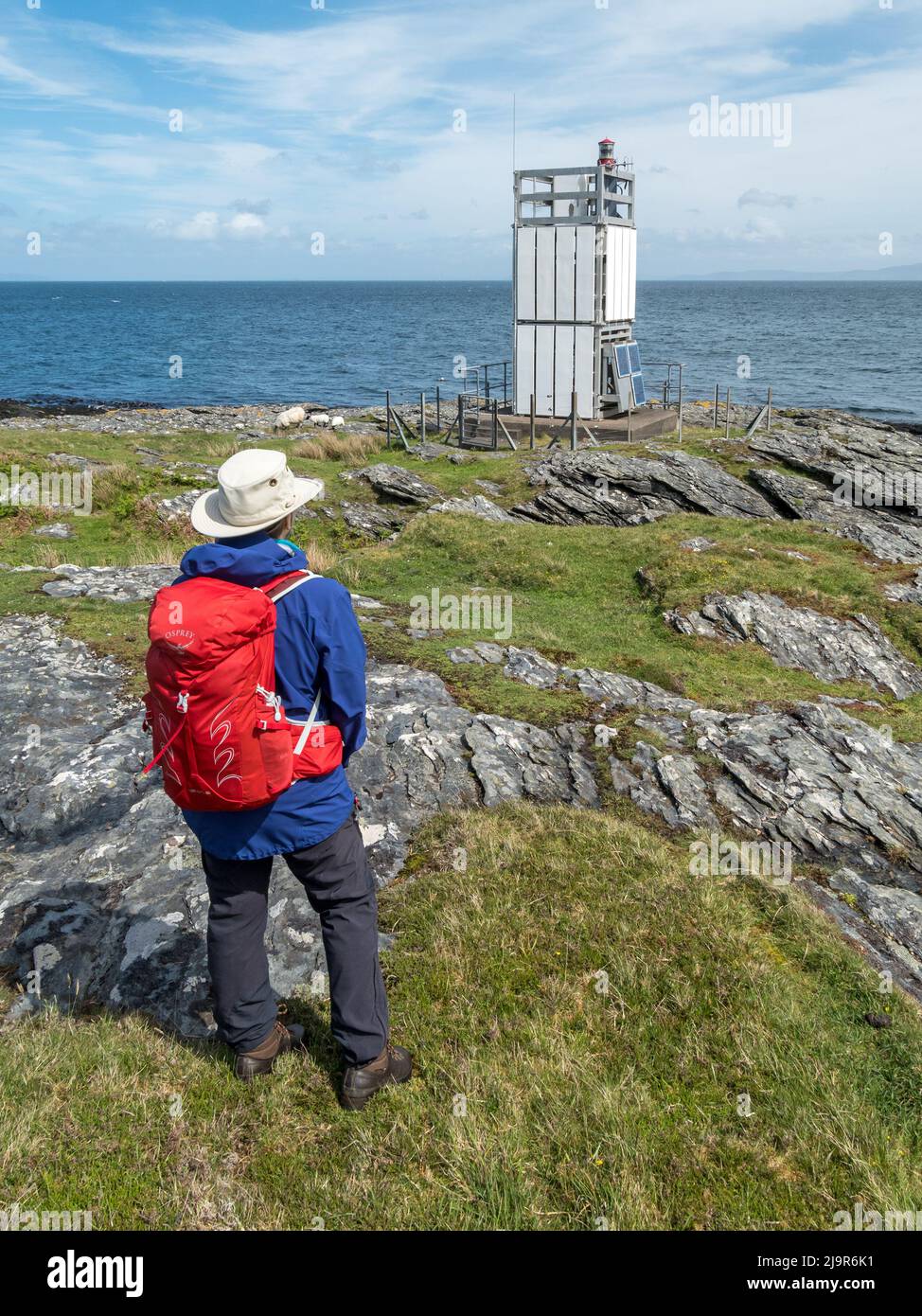 Female rambler looking out to sea and at the modern, small, unmanned solar powered lighthouse at Scalasaig on the Isle of Colonsay, Scotland, UK Stock Photo