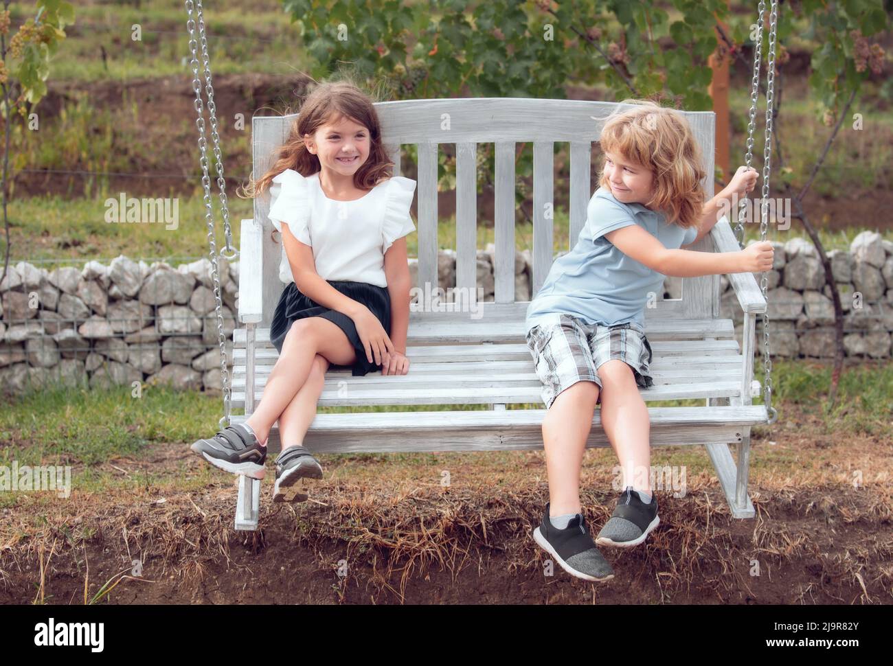 Happy brother and sister swinging on swing in summer park outdoors. Little boy and girl kids enjoying summer. Kids walking at backyard. Best friends Stock Photo