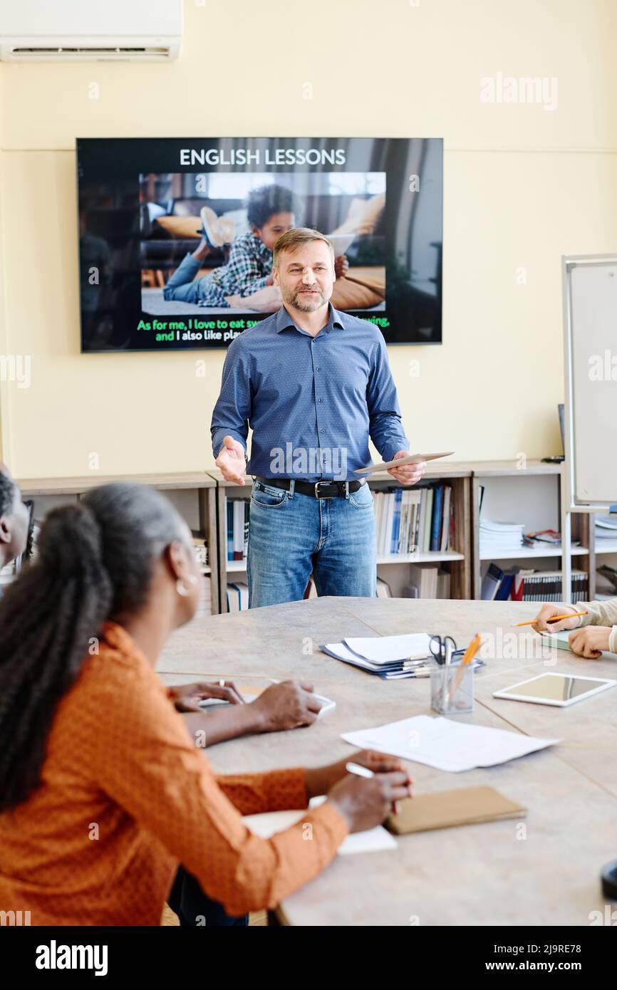 Vertical shot of mature Caucasian man standing at desk in front of multi-ethnic students teaching them English language Stock Photo