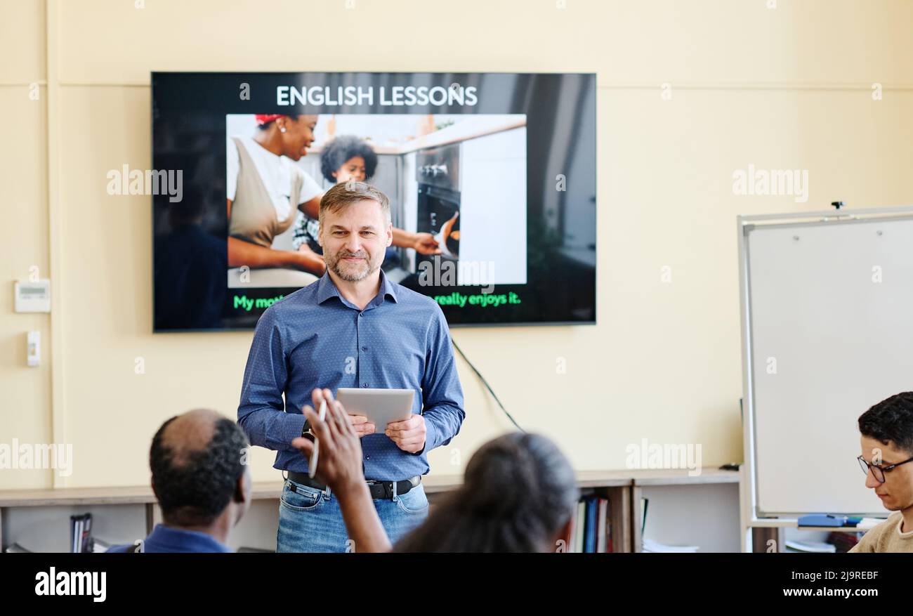 Mature Caucasian teacher holding digital tablet standing against screen with presentation on it working with migrant students Stock Photo