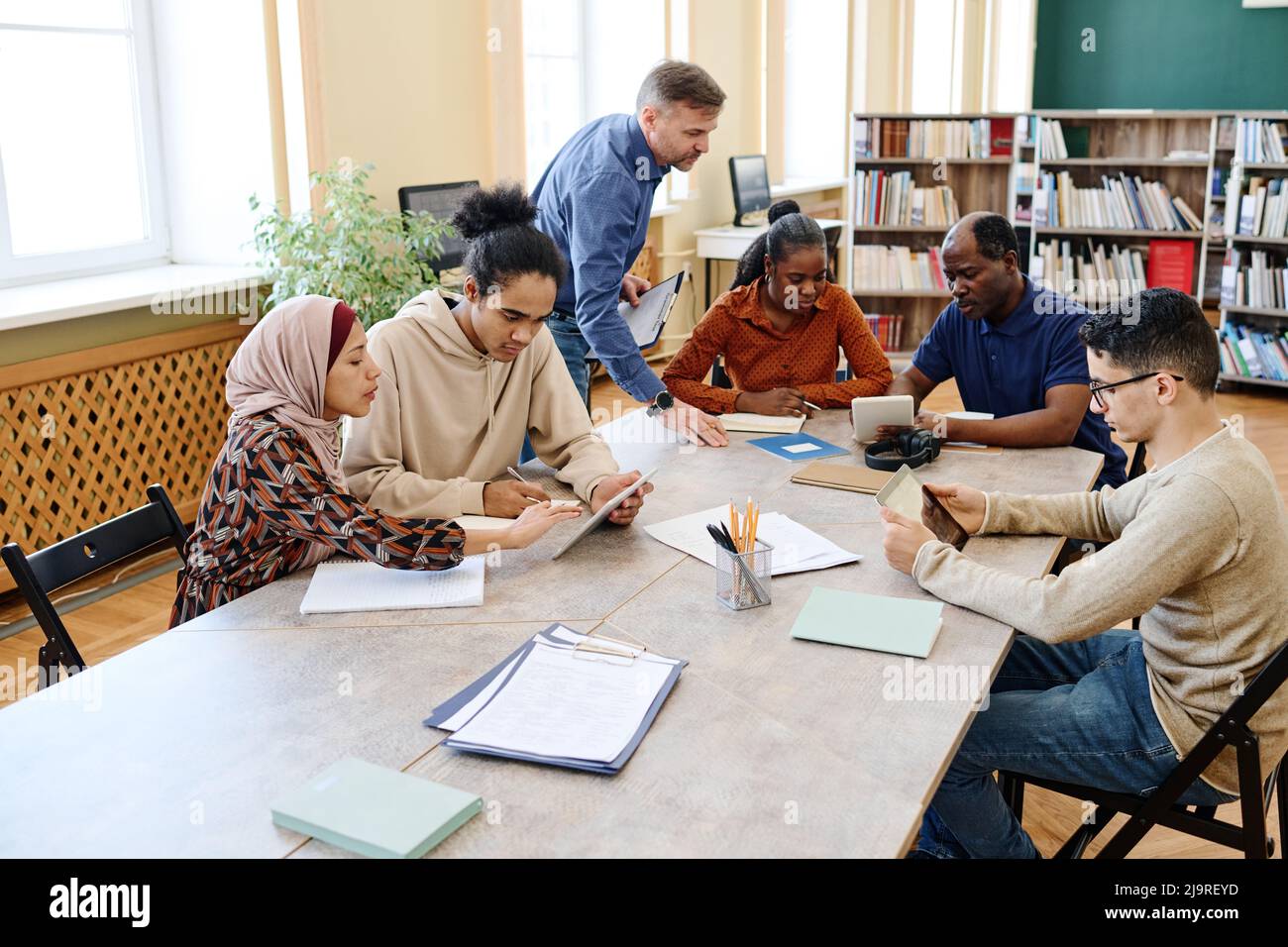 Mature Caucasian English language teacher helping immigrant students with doing task during lesson in library Stock Photo