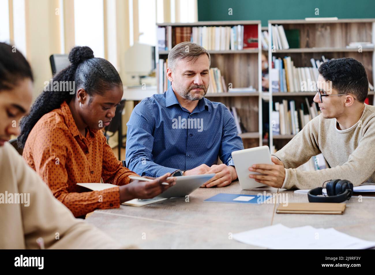 Mature language teacher sitting at table with his ethnically diverse students watching them doing exercise and answering questions Stock Photo
