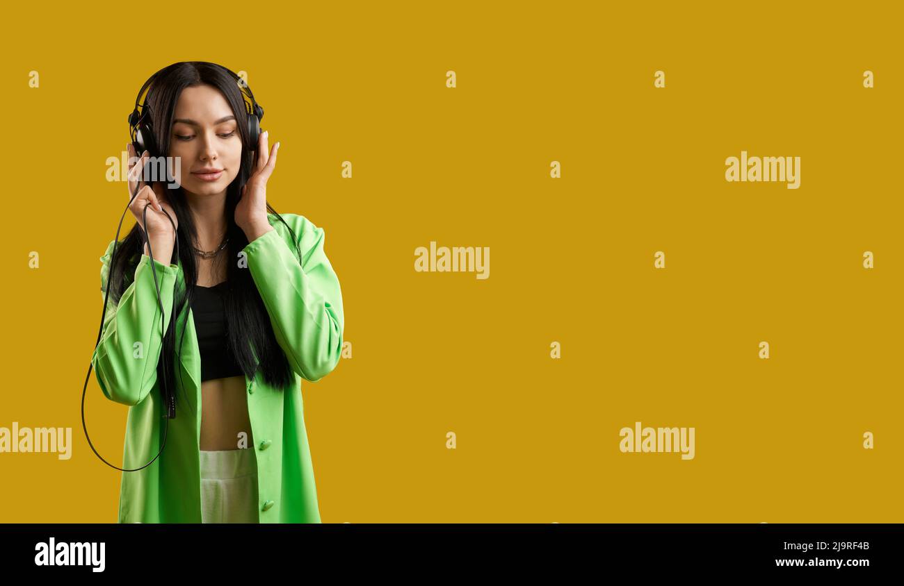 Front view of girl wearng green suit, standing, listening to music. Pretty brunette woman with long hair enjoying music with closed eyes, smiling. Concept of modern life. Stock Photo