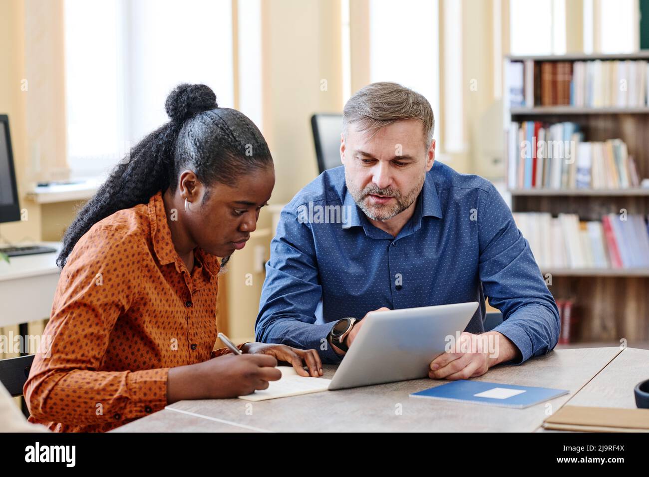 Mature Caucasian teacher sitting next to his Black student explaining her how to do task and showing something on digital tablet Stock Photo