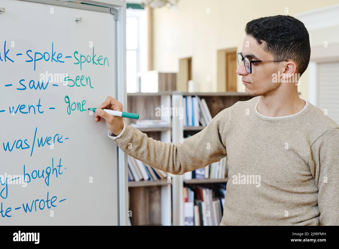 Handsome young Middle Eastern migrant student standing at whiteboard doing task with irregular verbs Stock Photo