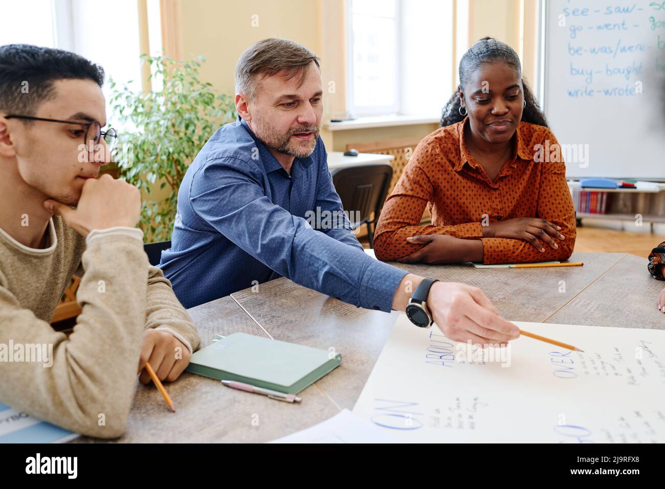Mature teacher working with immigrant students pointing pencil on word written on poster and explaining it Stock Photo