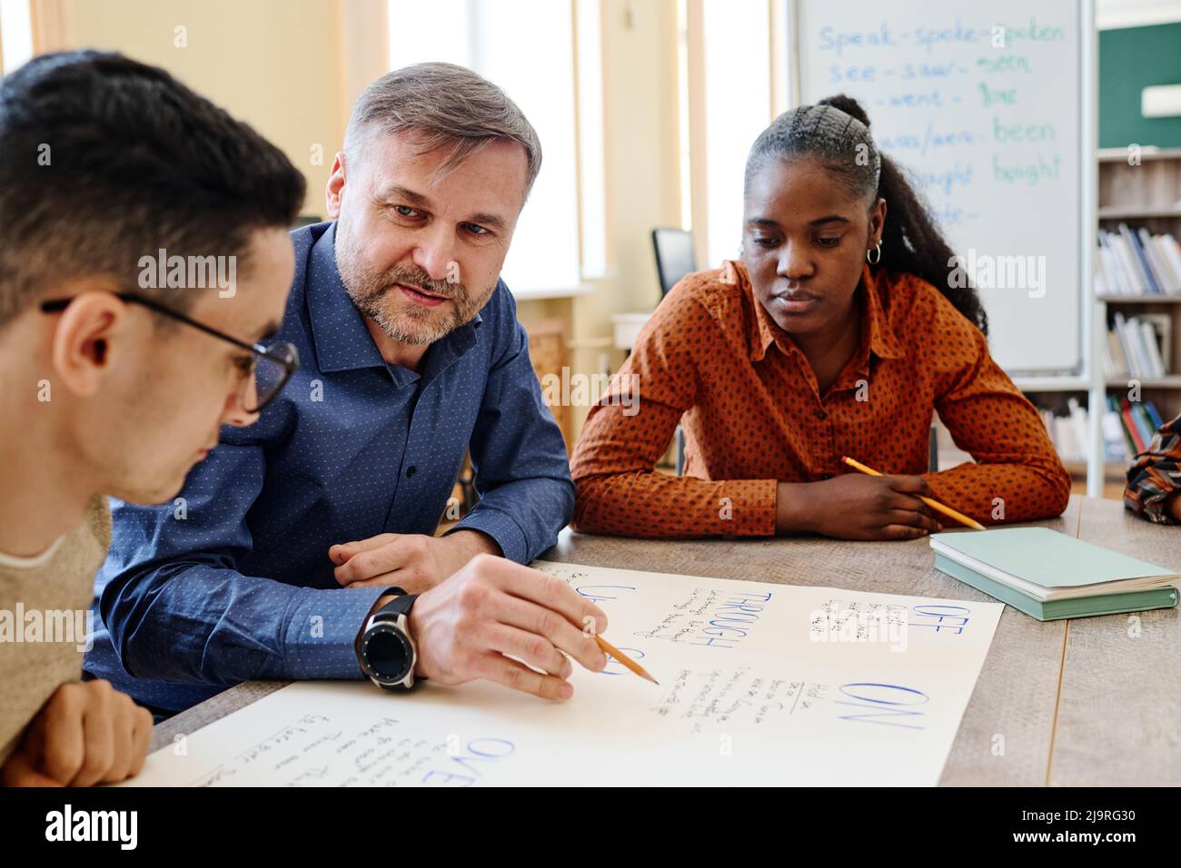 Mature English language teacher sitting at table with his students asking them questions during lesson Stock Photo