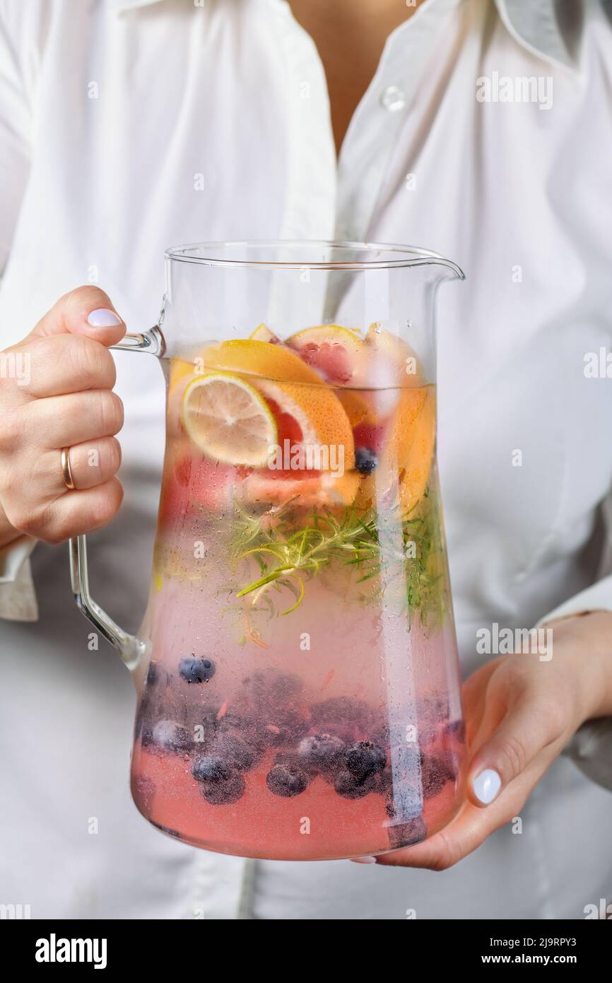 A jug with a refreshing organic soft drink with lime, blueberry, grapefruit and rosemary in female hands close-up Stock Photo