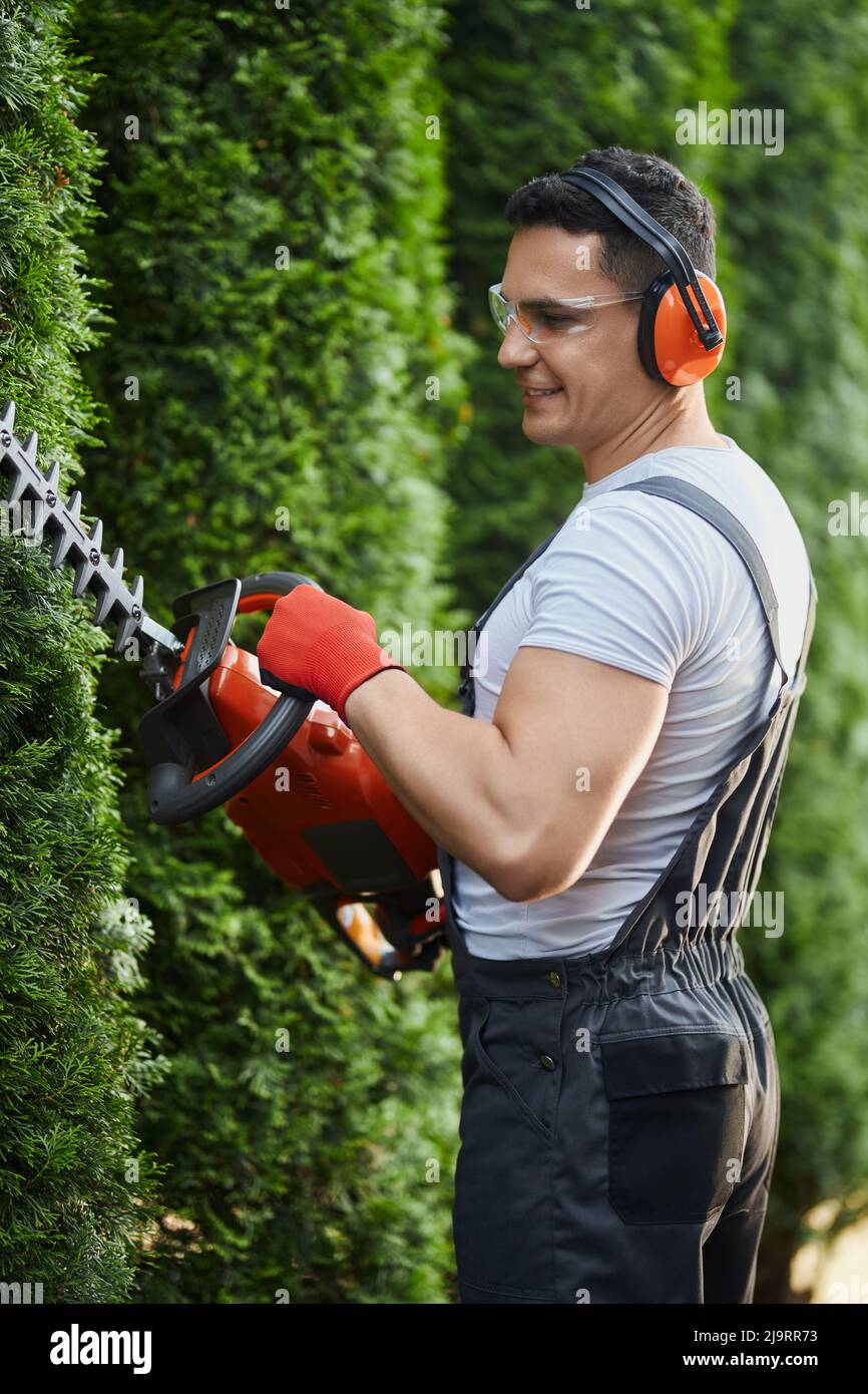Side view of happy caucasian man trimming hedge with electric cutter