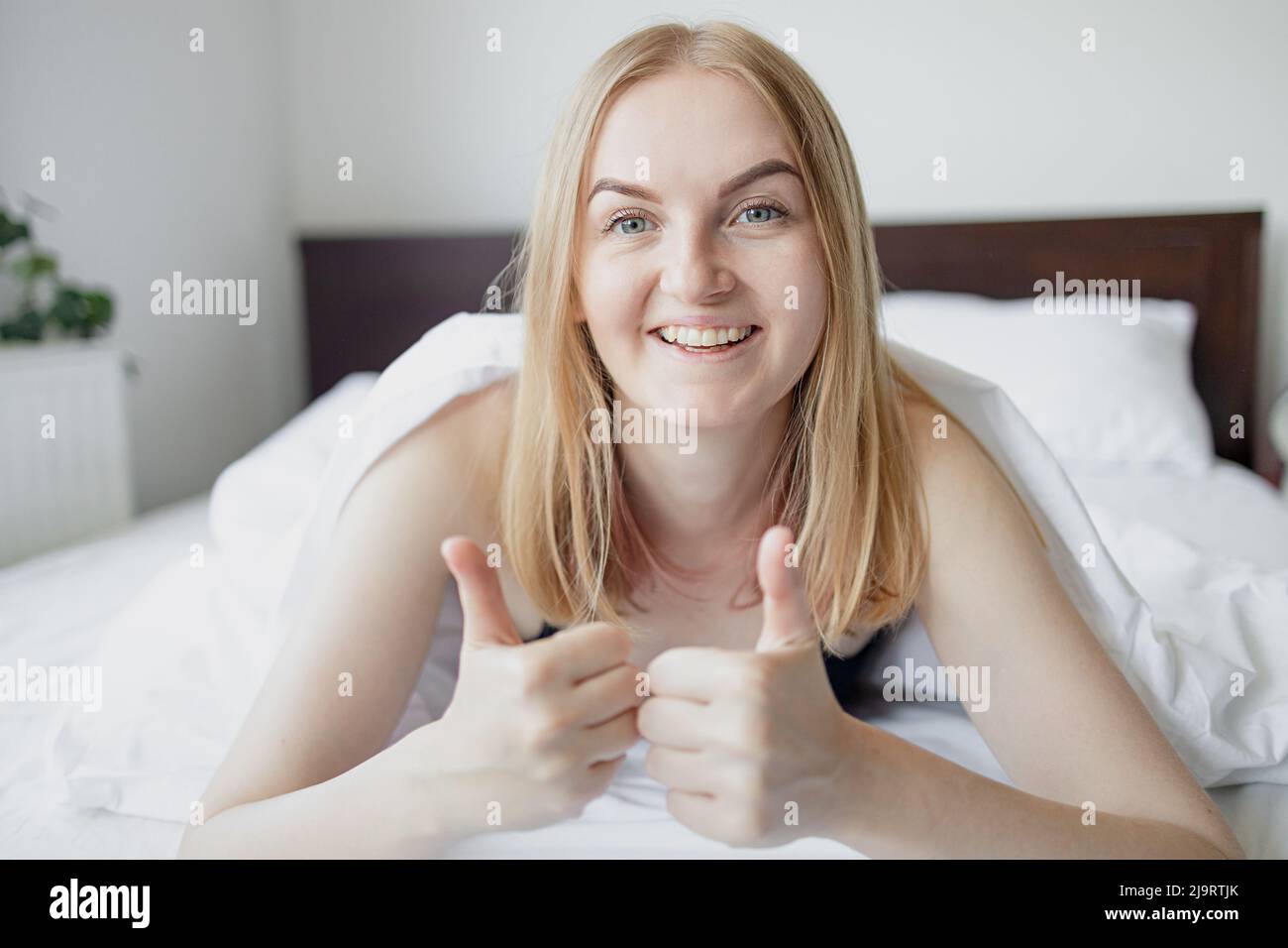 Happy young blonde 30s woman shows like with hand laying in bed covered with white bedclothes early in the morning.  Stock Photo