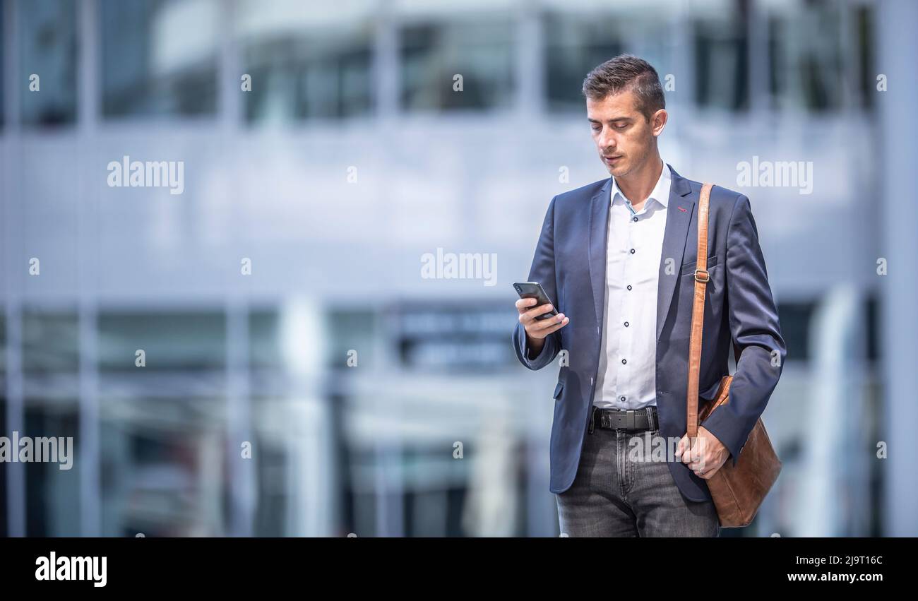Businessman in a casual outfit wears mens' purse, standing in front of an office building, chekcing his cell phone. Stock Photo
