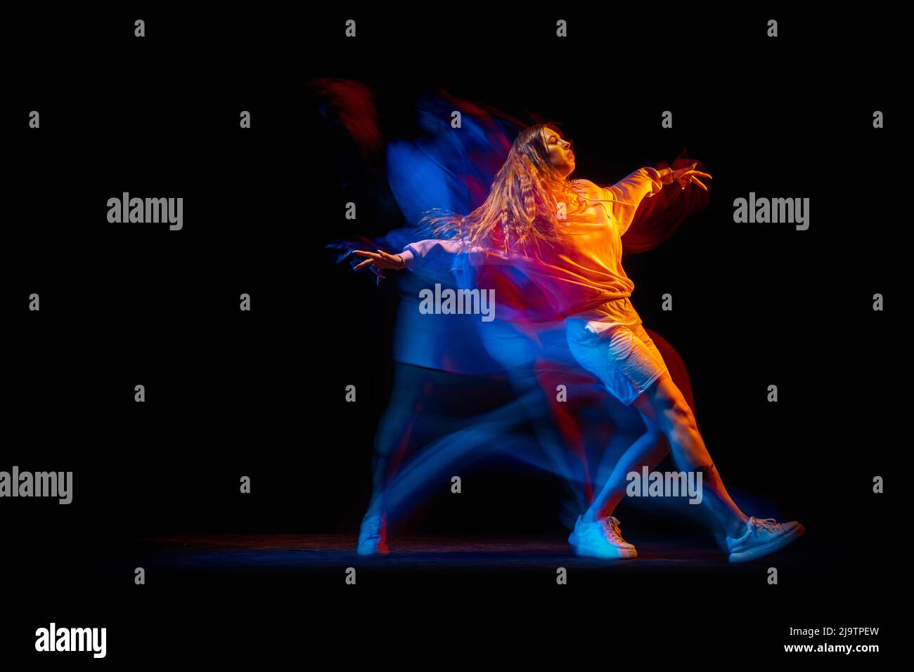 Young woman dancing hip-hop in sportive style clothes isolated on dark background at dance hall in mixed neon light. Youth culture, hip-hop, movement Stock Photo