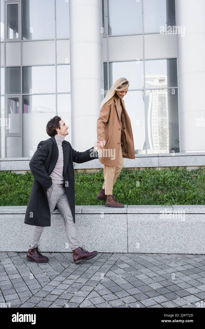 full length of man with hand in pocket holding hand of woman walking on