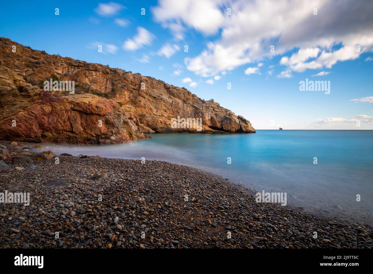 Photograph taken with long exposure on a beach with crystal blue waters in Cartagena, in the region of Murcia, Spain on a sunny day, with boulders on Stock Photo