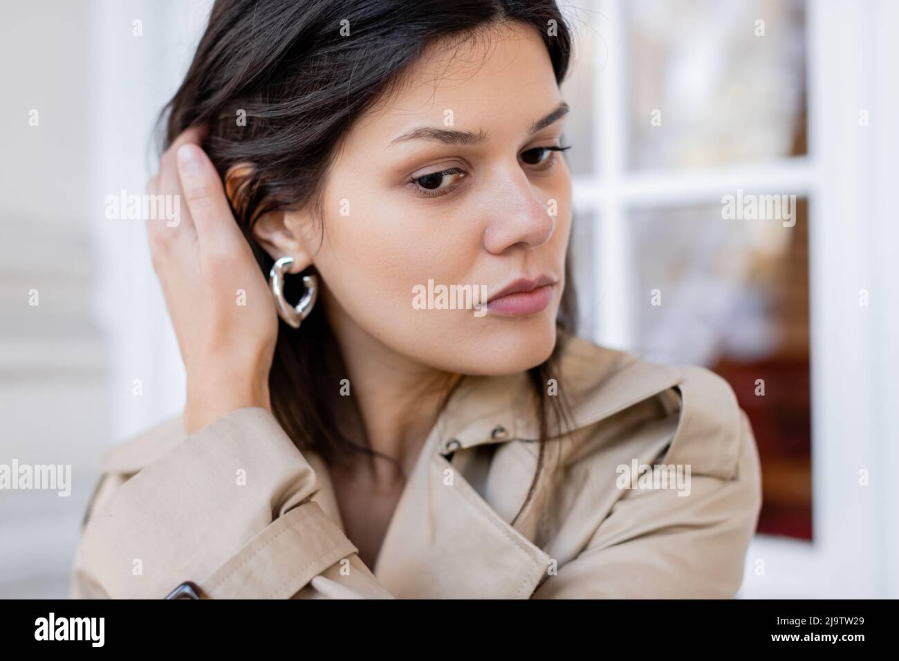 portrait of brunette woman in beige trench coat and hoop earring adjusting hair outside Stock Photo
