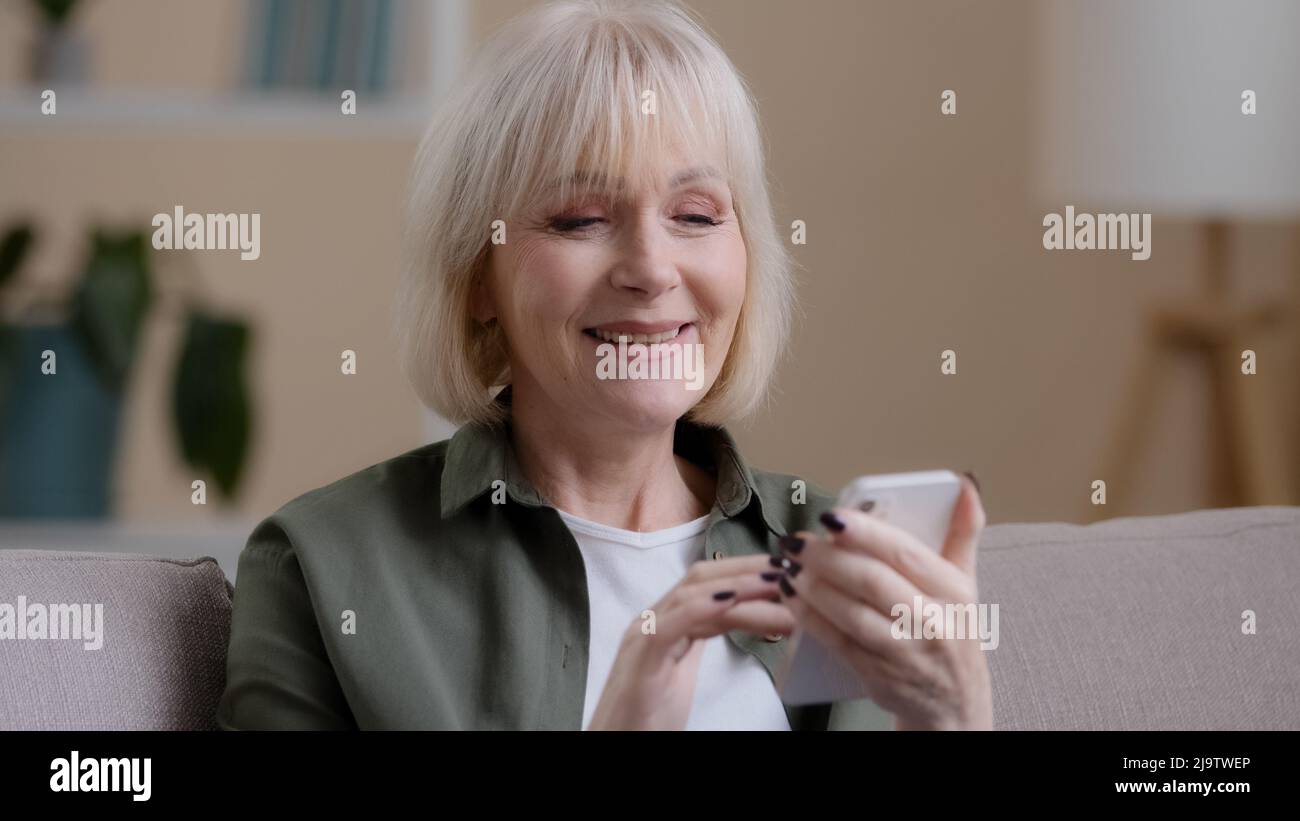 Mature elderly mature 60s caucasian woman with phone female smiling uses mobile app at home buys medicine online choosing clothes internet shopping e Stock Photo