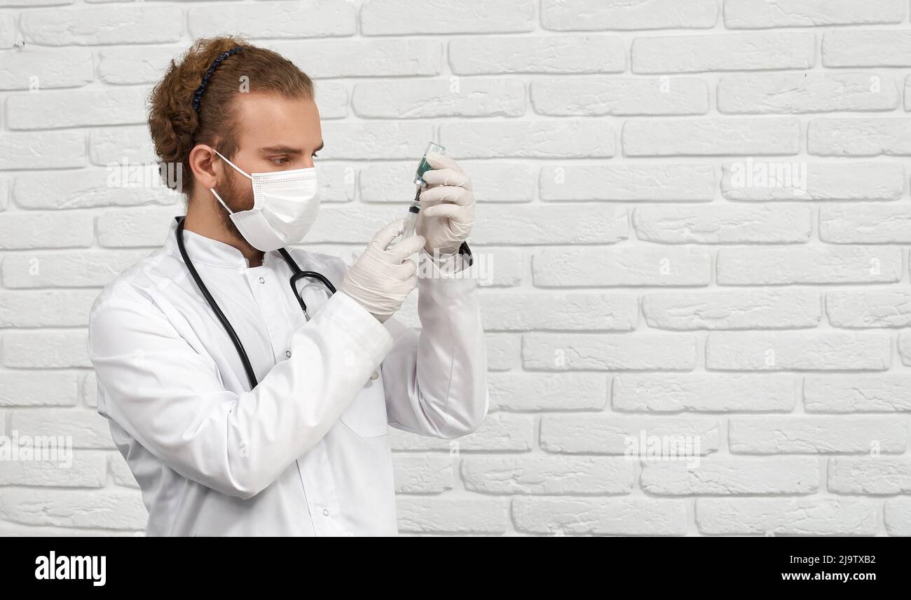 Side view of male vet in mask lab coat and gloves filling syringe with medication before injection. Crop of medical worker gaining medicine from vial on white brick wall background. Concept of treat. Stock Photo
