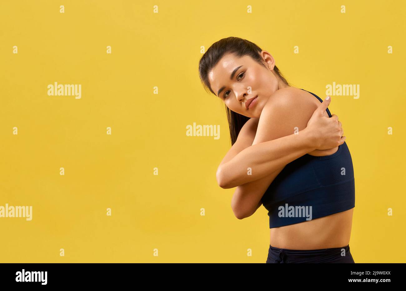 Young woman in black top tight hugging herself, thoughtfully looking at camera. Portrait view of serious girl wrapping arms around body in hug, isolated on orange background. Concept of mindfulness. Stock Photo