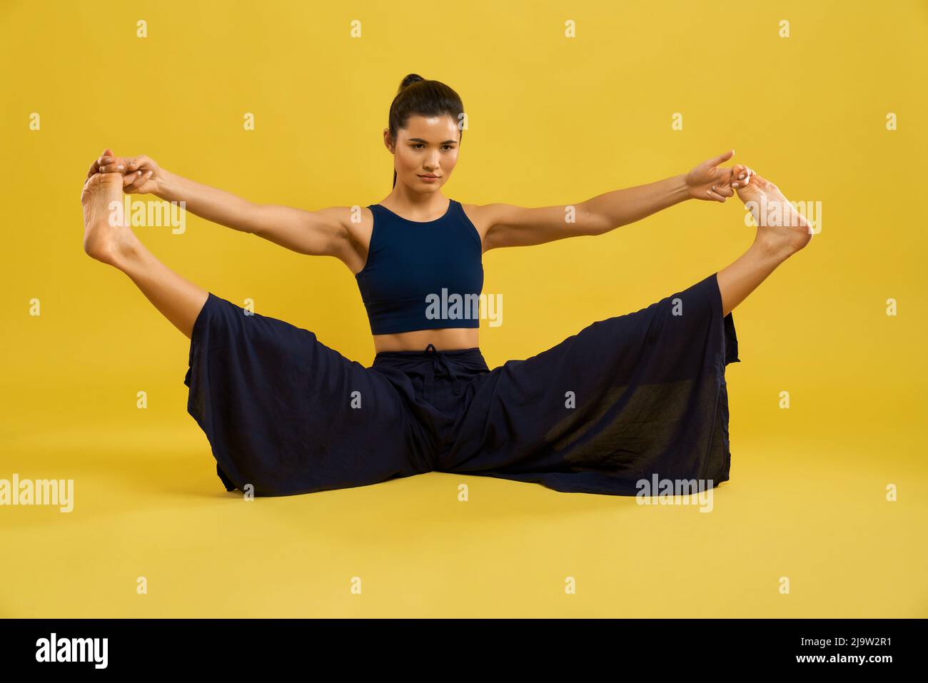 Fit caucasian girl stretching legs in Upavistha Konasana during yoga training. Front view of brunette woman in wide pants practicing yoga, isolated on tangerine studio background. Concept of yoga. Stock Photo