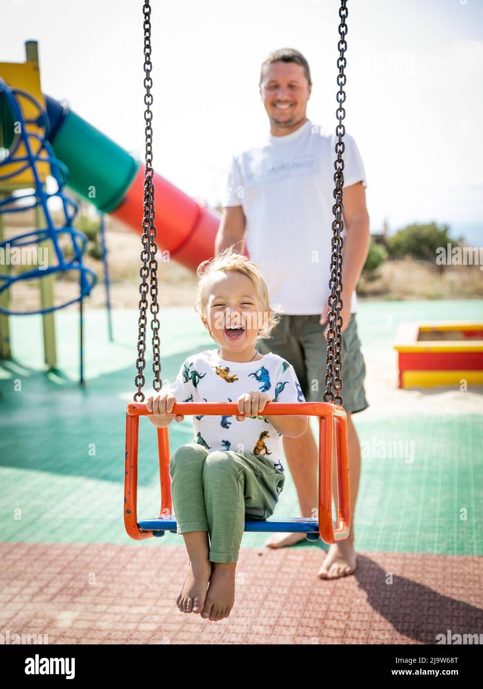 Young father enjoys parenthood with his little son barefoot in the playground on a sunny warm summer day. The father swings the baby on the swing and Stock Photo