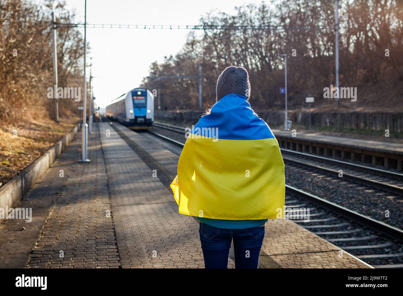 Woman wrapped in ukrainian flag standing at railroad station platform and looking to arriving train. Concept of humanitarian crisis and leaving refuge Stock Photo