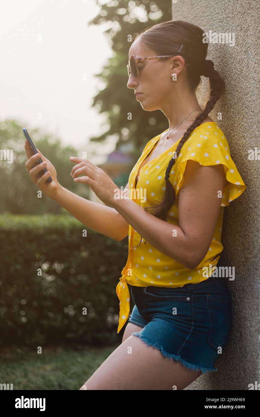 young woman on summer vacation with short jeans and yellow t-shirt with white dots and sunglasses at sunset sending messages from her mobile phone. Ou Stock Photo