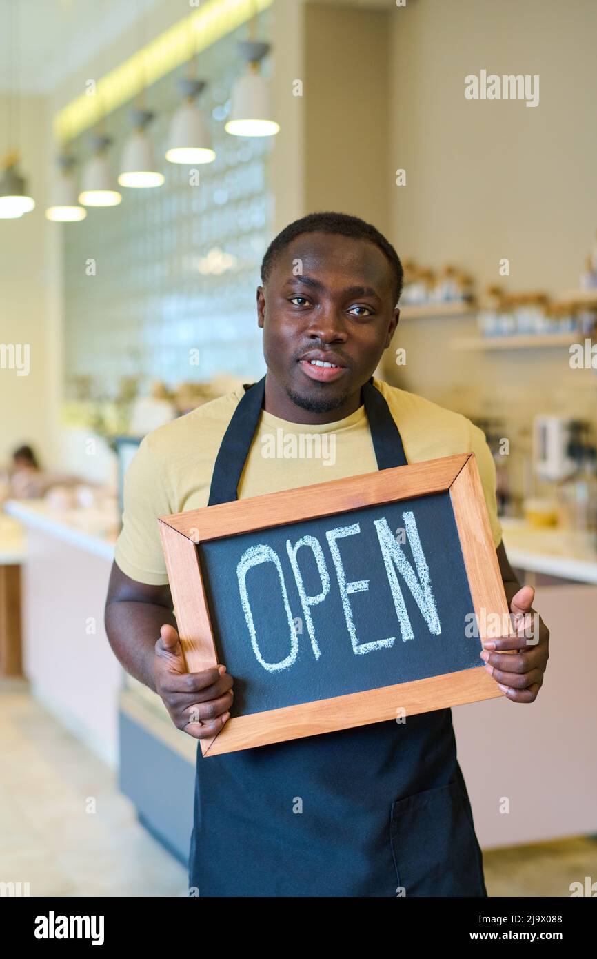 Portrait of African young waiter in uniform holding open sign and looking at camera, he opening new coffee shop Stock Photo