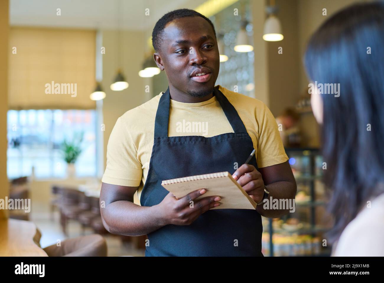 African American waiter making notes in notepad while talking to woman in cafe, he taking order from her Stock Photo