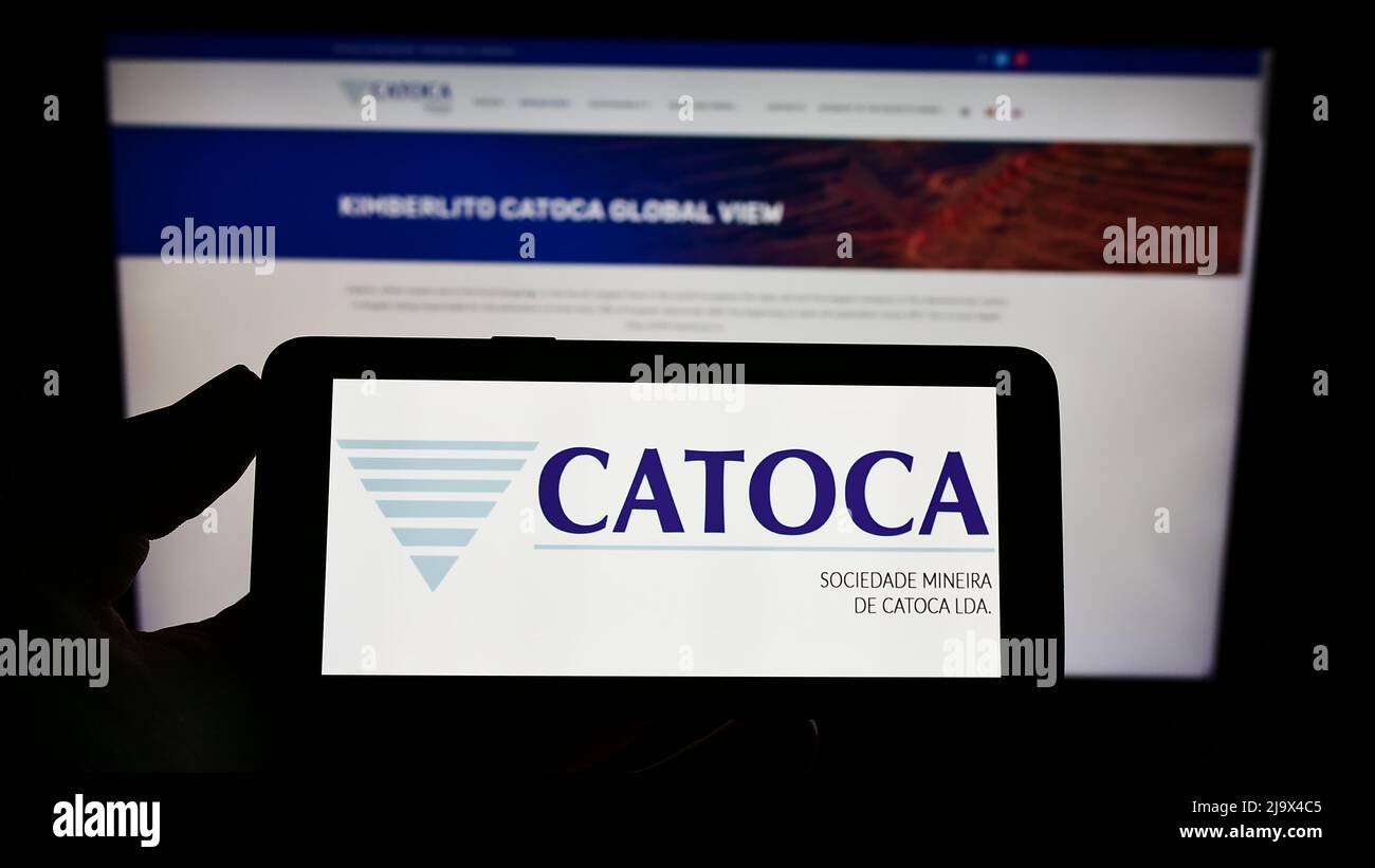 Person holding mobile phone with logo of mining company Sociedade Mineira de Catoca Lda. on screen in front of web page. Focus on phone display. Stock Photo