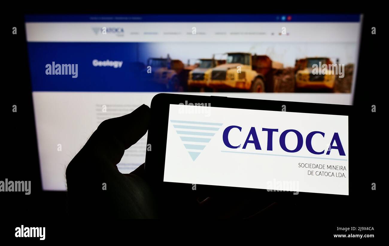 Person holding cellphone with logo of mining company Sociedade Mineira de Catoca Lda. on screen in front of webpage. Focus on phone display. Stock Photo