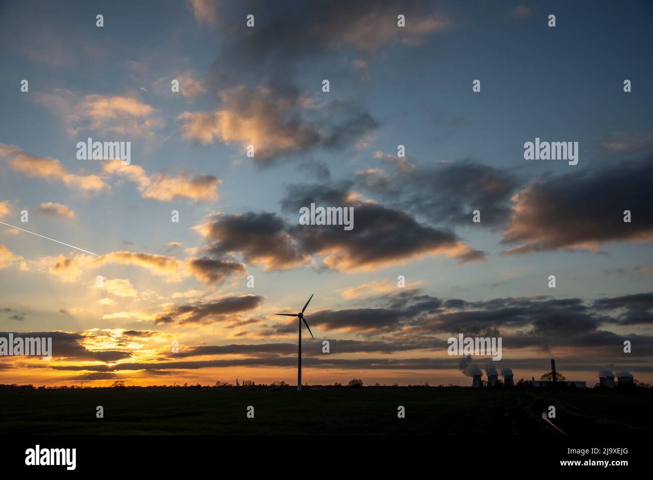 A wind turbine silhouetted against the setting sun, with the cooling towers of a power station in the background Stock Photo