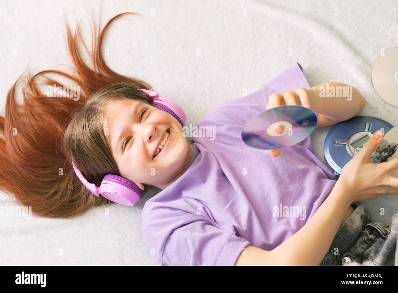 A happy teenage girl in a lilac T-shirt lies on the bed headphones and listens to music on CD player Stock Photo