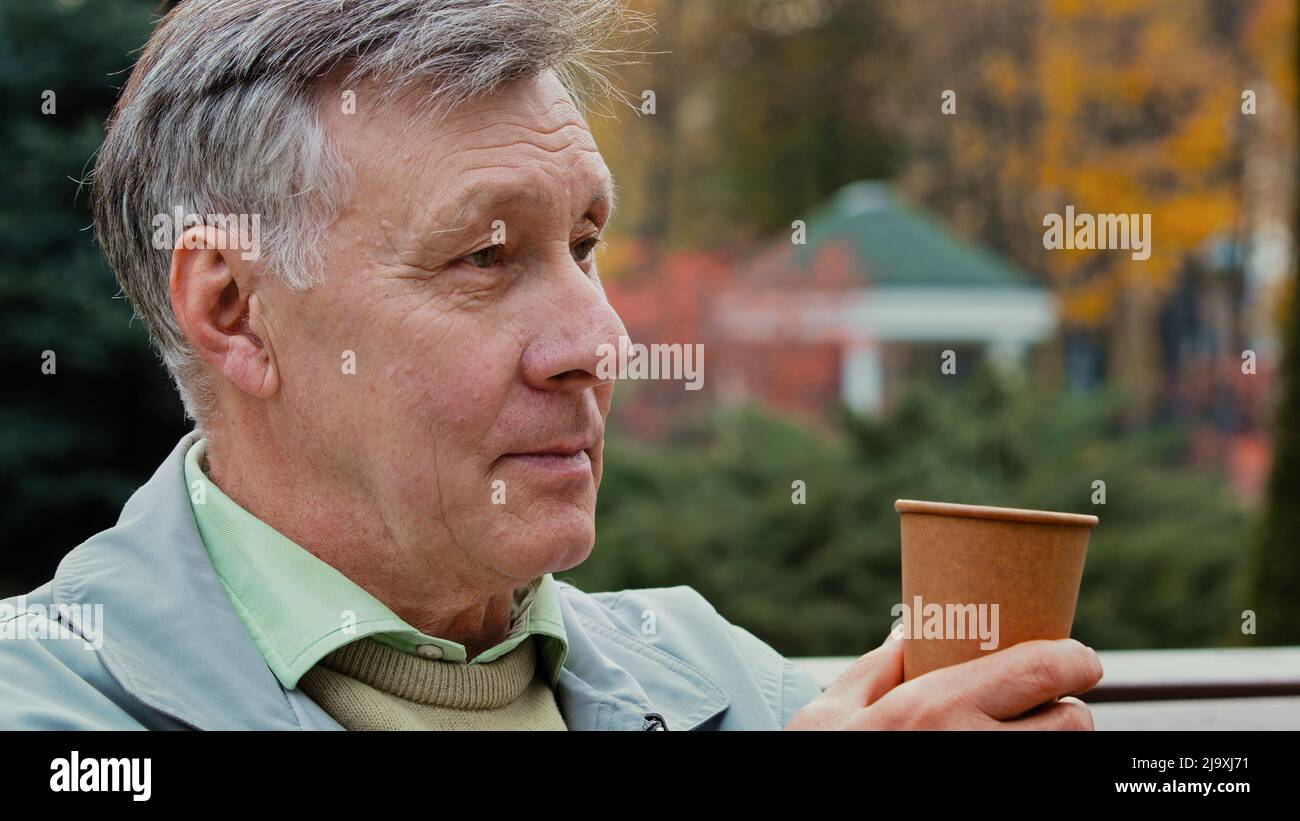 Dreamy calm gray-haired elderly pensioner man grandfather middle aged adult male person sitting on autumn park bench drinking coffee hot tea Stock Photo