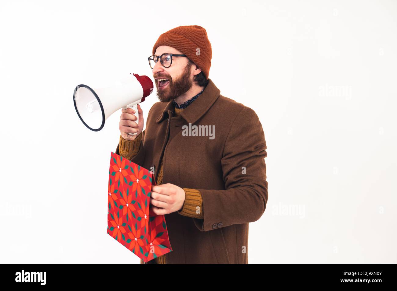 Caucasian man with a megaphone. Studio shot of a millennial bearded guy in a beanie and brown coat holding a loudspeaker and a gift bag over white background,. High quality photo Stock Photo