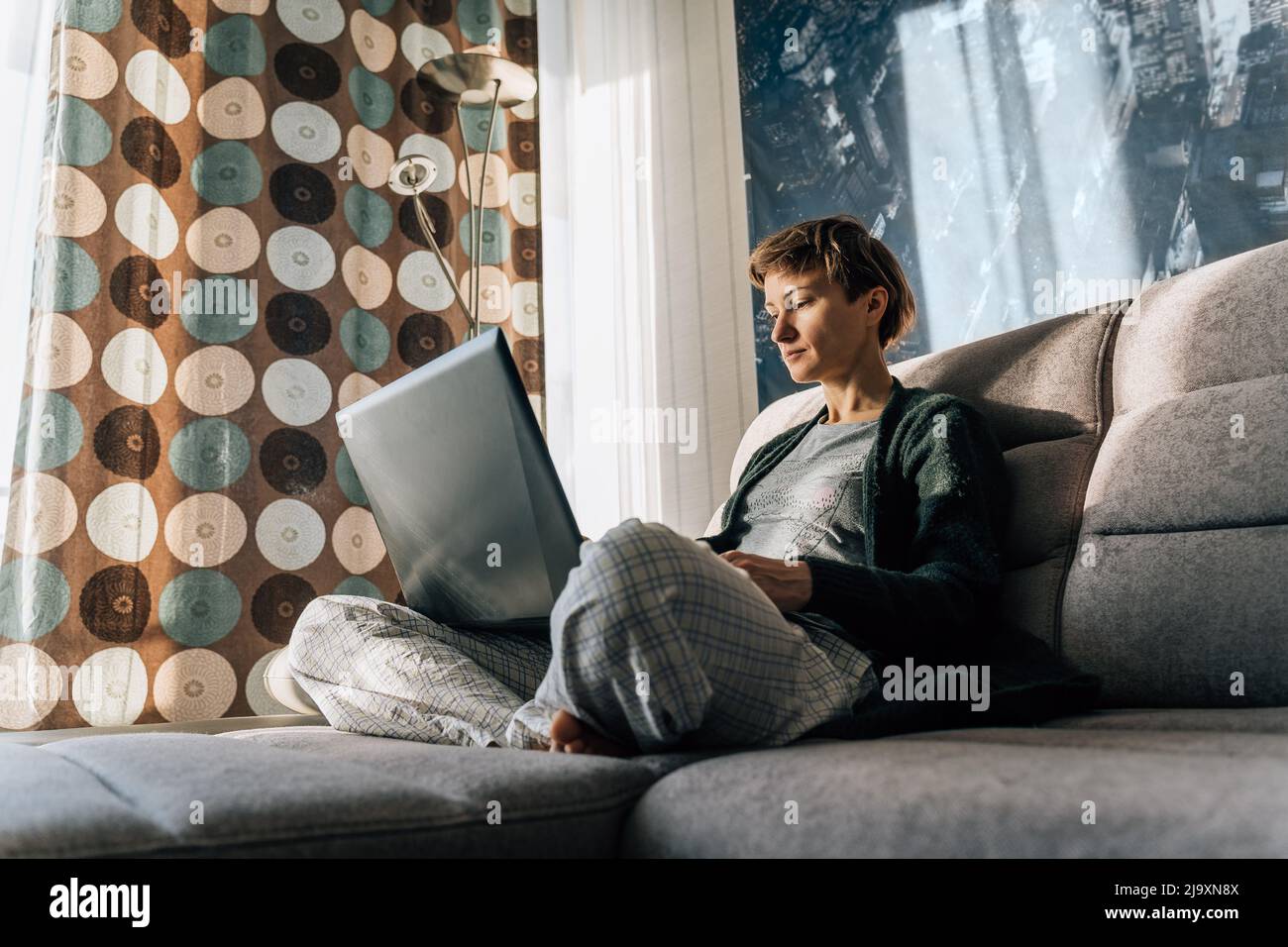 A woman sits comfortably on the couch, legs crossed, and uses a laptop Stock Photo
