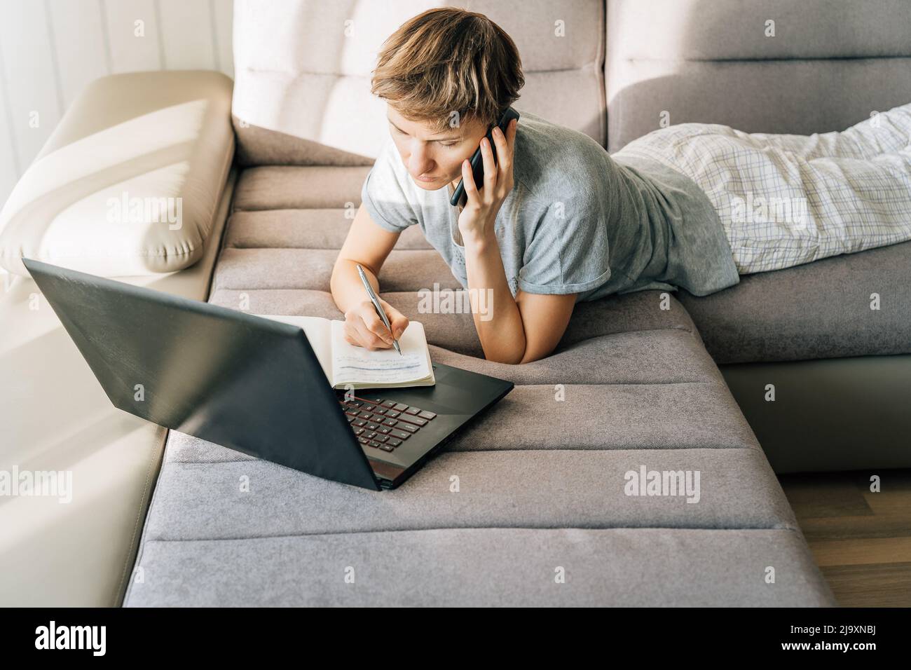 Woman lies on couch, talking on the phone and taking notes Stock Photo