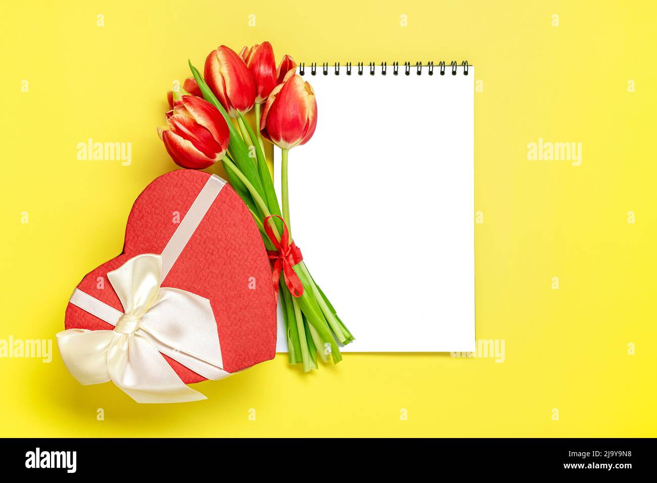 Bouquet of red tulips, gift box heart, notepad on yellow background Top view Flat lay Holiday greeting card Happy moter's day, 8 March, Valentine's day, Easter concept Copy space Mock up. Stock Photo