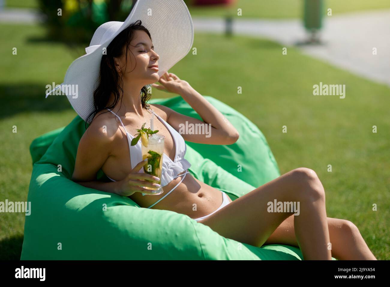 Relaxed woman in white bikini resting with fresh cocktail at grassy area of resort. Front view beautiful lady in lovely hat enjoying mojito, while chilling on bag chair outdoors. Concept of vacation. Stock Photo