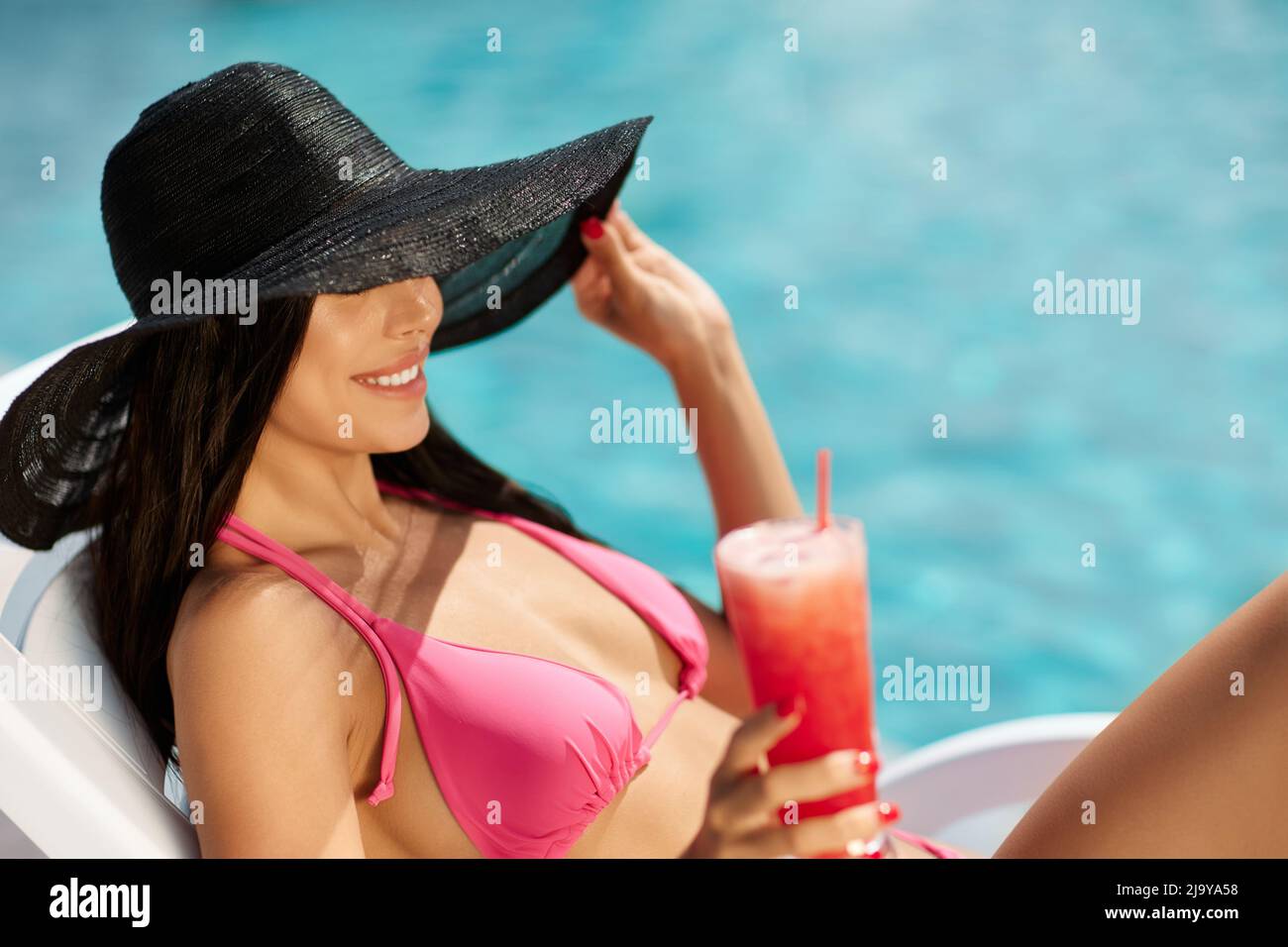 Pretty brunette resting on lounger, hiding eyes under brim of black hat outdoors. Side view of charming female in pink bikini drinking fresh smoothie, while lying on sunbed at resort. Concept of rest. Stock Photo