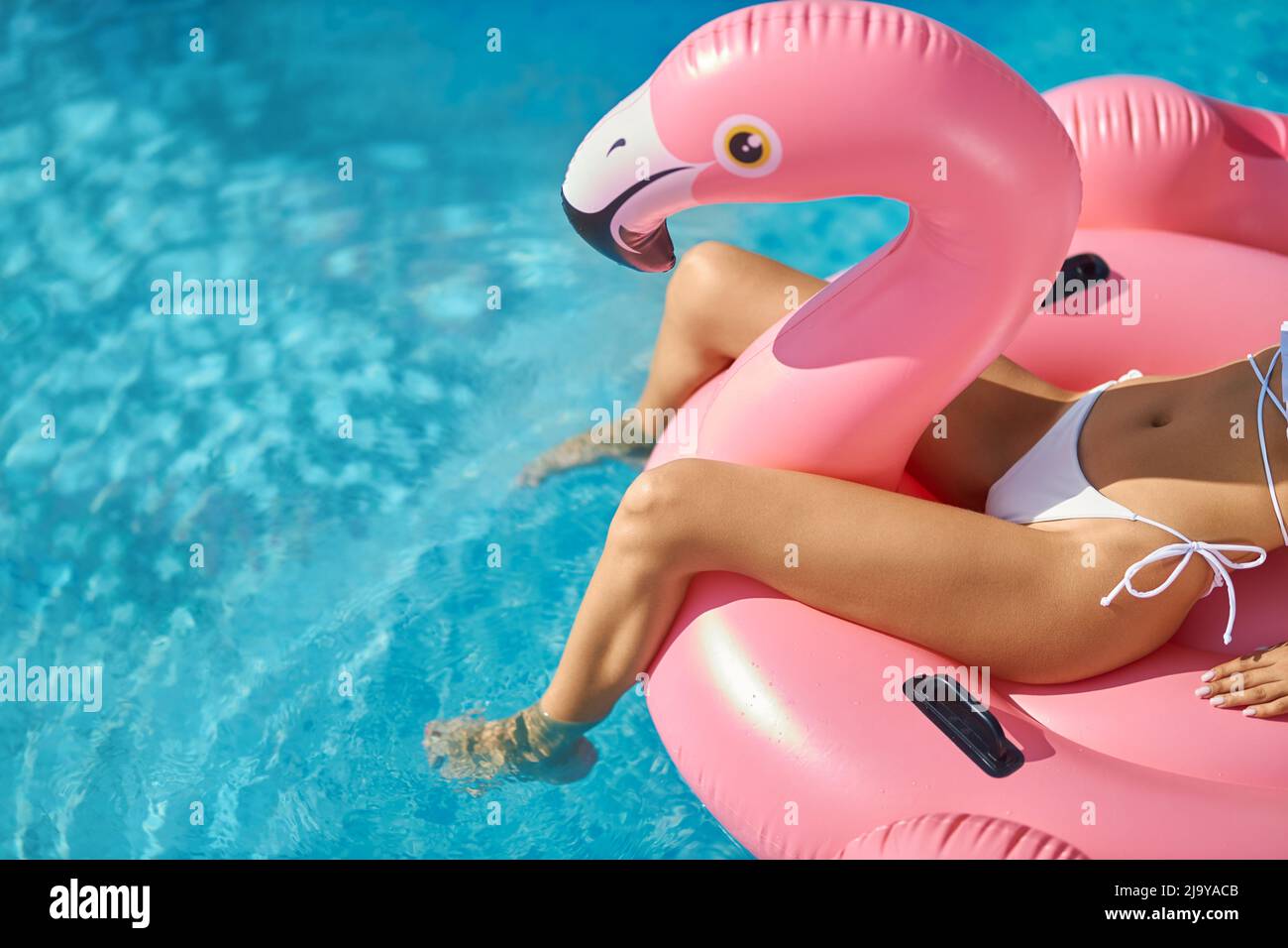 Beautiful tanned female relaxing on coral flamingo bird in swimming pool. Crop view of slim woman in white bikini sunbathing, while floating on blue clear water surface at hot day. Concept of relax. Stock Photo