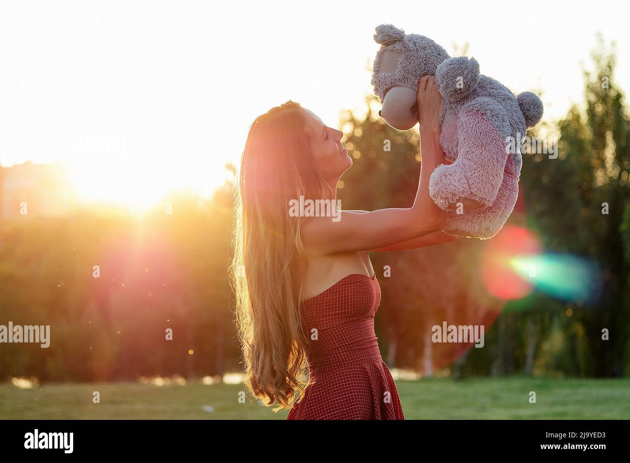 cute long-haired tanned woman enigmatic smile in a red dress holding a gray teddy bear toy in hands in the park trees background Stock Photo
