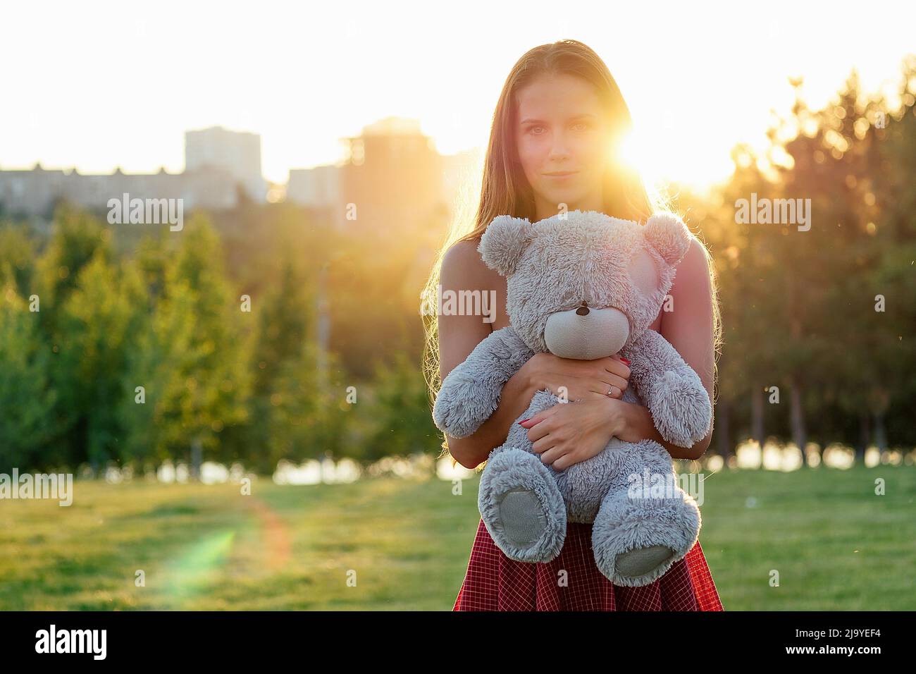 cute long-haired tanned girl enigmatic smile holding a gray teddy bear toy in hands in the park trees background Stock Photo