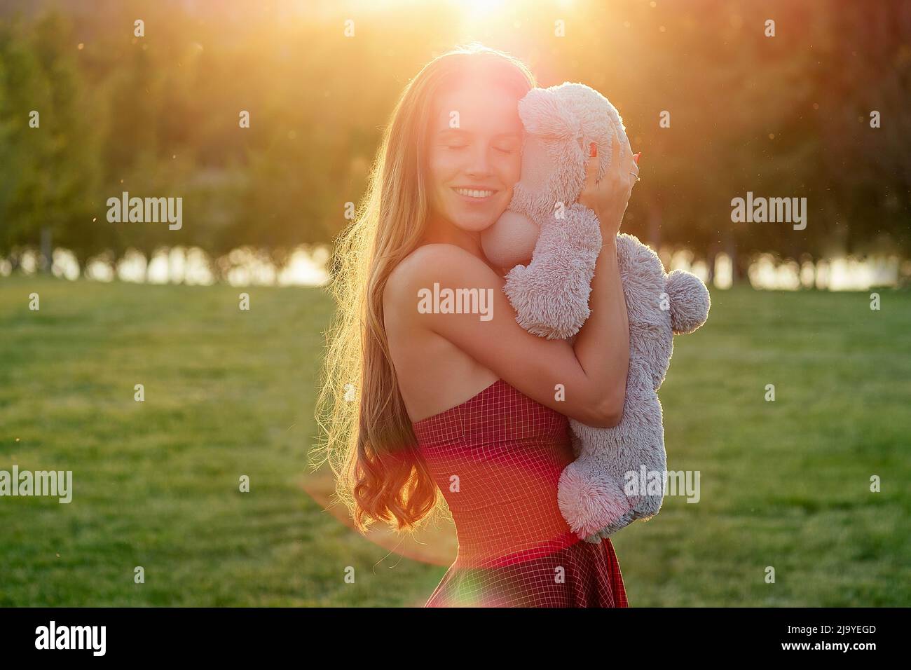 cute long-haired tanned woman enigmatic smile in a red dress holding a gray teddy bear toy in hands in the park trees background Stock Photo