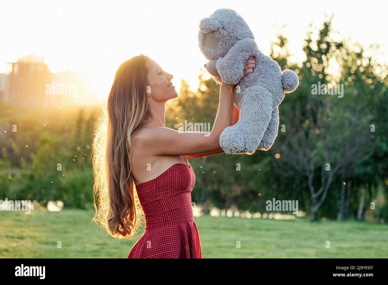 cute long-haired tanned woman enigmatic smile holding a gray teddy bear toy in hands in the park trees background Stock Photo