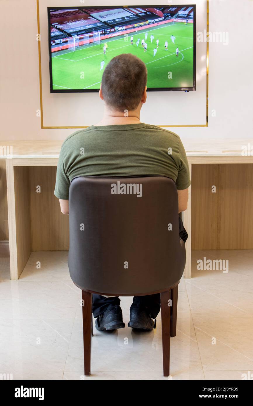 A lone man sits uncomfortably in a chair and watches a football match on television hanging on the wall of the room. Stock Photo