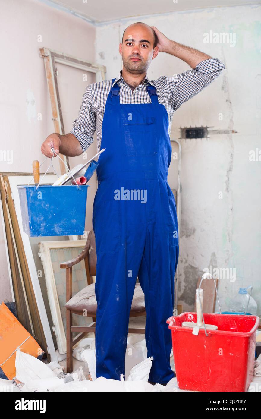 Builder is standing with bucket and putty knife and thinking about where he should start Stock Photo