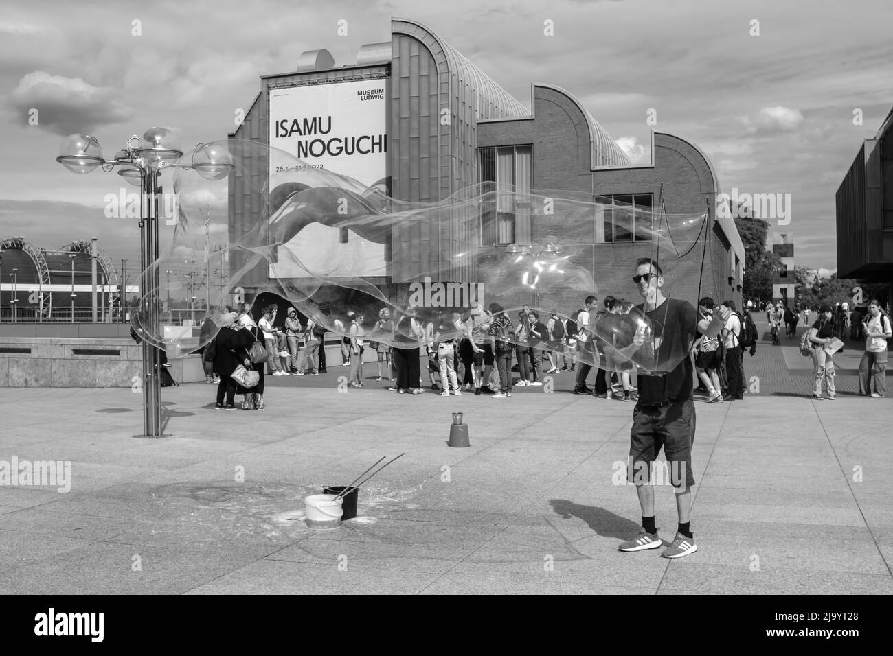 Cologne, Germany - May 17, 2022 : A street performer making gigantic soap bubbles in front of a young crowd of people in black and white Stock Photo