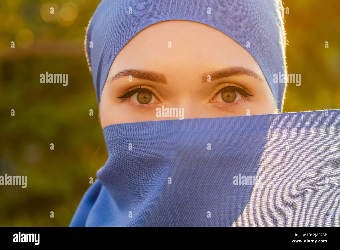 Portrait of beautiful Muslim woman with green eyes wearing blue scarf face closed covered with aveil background forest trees in the park Stock Photo